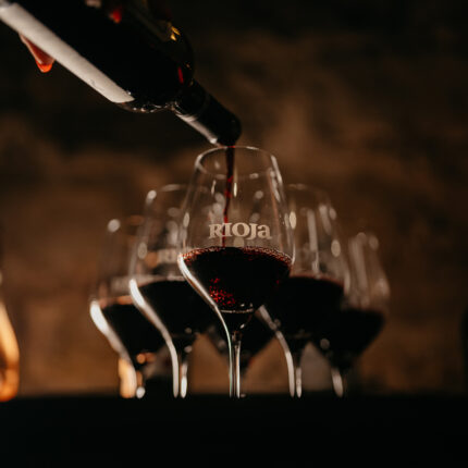 Red wine being poured from a bottle into a row of wine glasses, with the central glass.